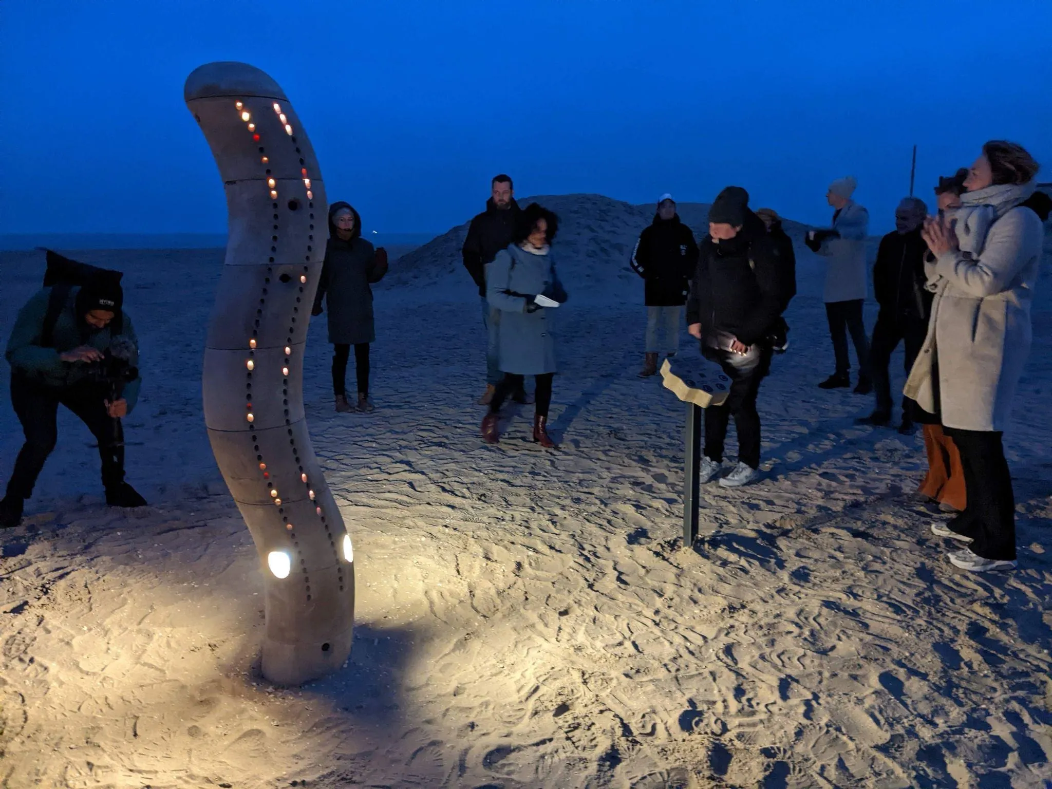 The Wave lit at dusk on Scheveningen beach, with a group of people gathered around it
